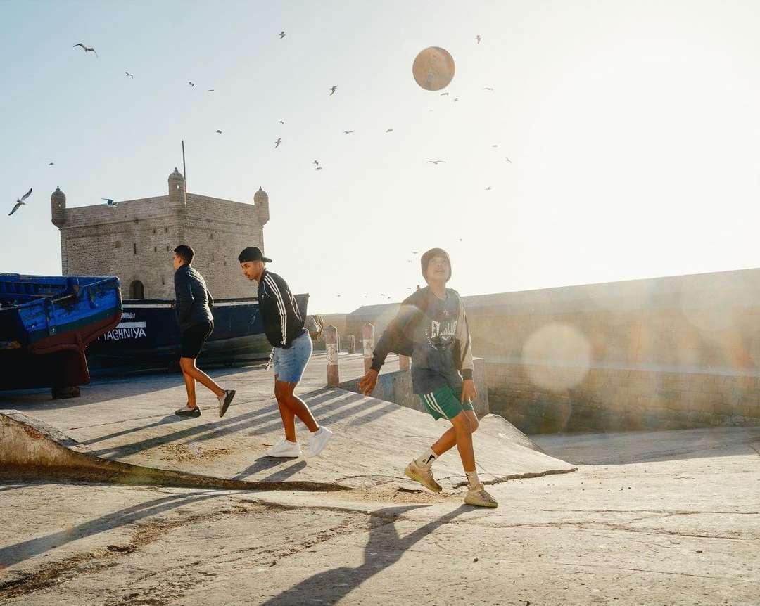 Three boys play with a soccer ball near Essaouira Citadel in Morocco with seagulls flying overhead on a bright, sunny day.
