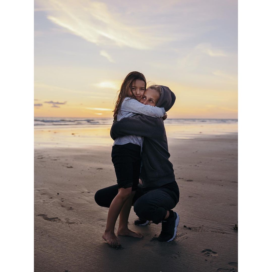 A warm embrace on the beach at sunset: A young girl hugs an adult on the sandy shore with gentle waves and a soft, colorful sky.