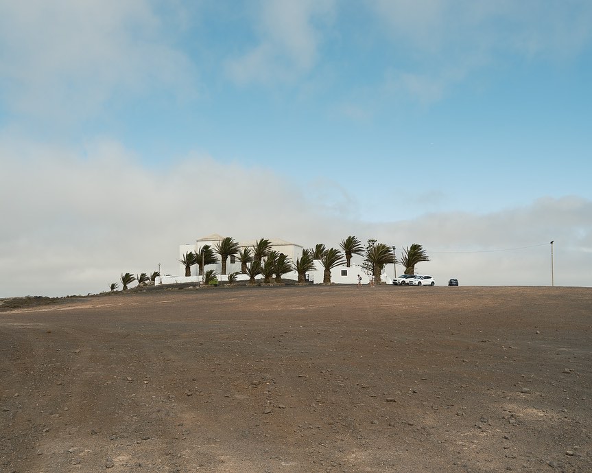 White building on a hill surrounded by palm trees under a cloudy sky, vehicles parked in front.