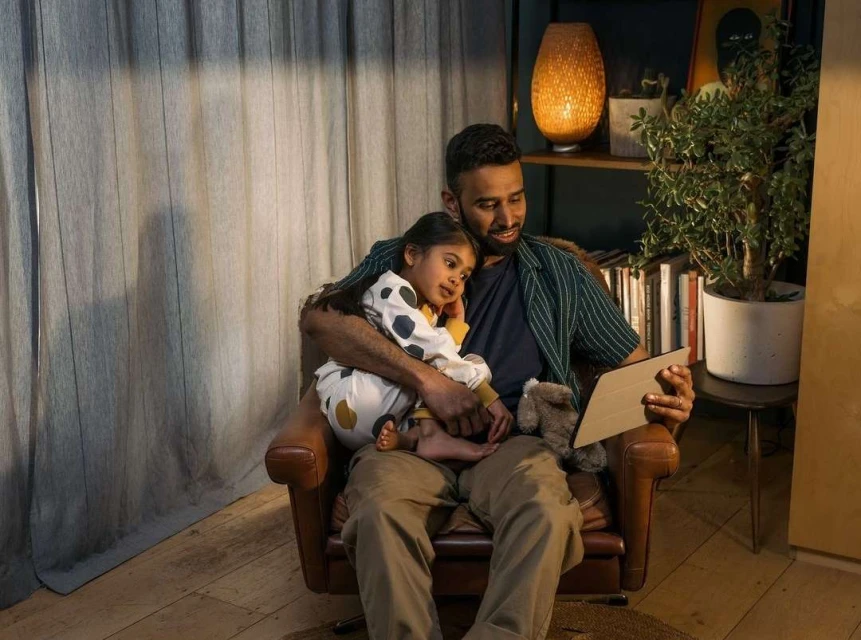 Father and daughter looking at a tablet in a warmly lit living room. The man and child are sitting in a brown leather chair.