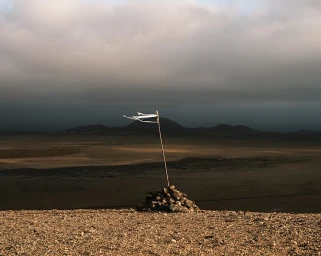 Tattered white fabric flag on a thin wooden pole atop a rock cairn overlooking a vast desert landscape under an overcast sky.