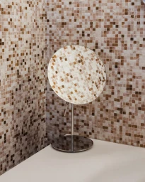 Round mosaic vanity mirror on a thin metal stand against a matching brown and white tiled corner wall