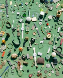 Overhead arrangement of vintage trinkets, wristwatches, and small collectibles spread across a green fabric surface