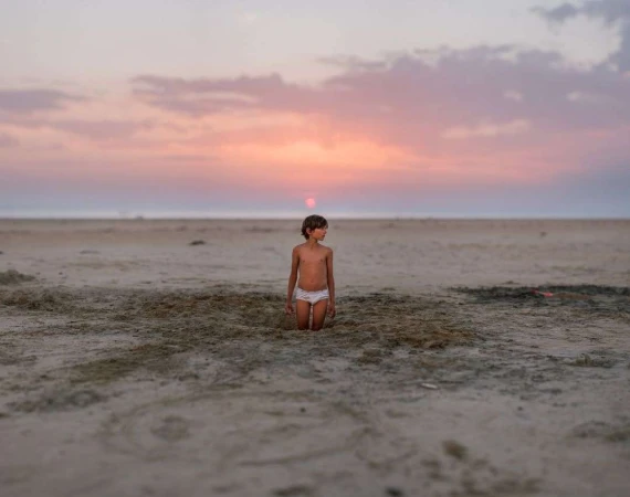 Young child standing in a sandy hole on a beach at sunset with a pink sky.