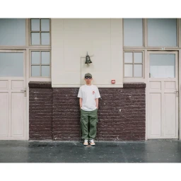 Man wearing a white t-shirt and green cargo trousers standing centrally against a brick and cream wall with double doors.