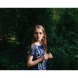 Young girl with long blonde hair wearing a blue graphic t-shirt standing outdoors in a lush forest by Alex Cretey Systermans