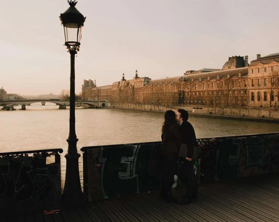 A couple kissing on a bridge by a river with grand stone buildings along the opposite bank at sunset.