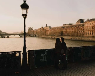 A couple kissing on a bridge by a river with grand stone buildings along the opposite bank at sunset.