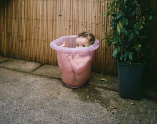 Baby sitting inside a pink plastic basin outdoors on a concrete ground next to a potted plant.