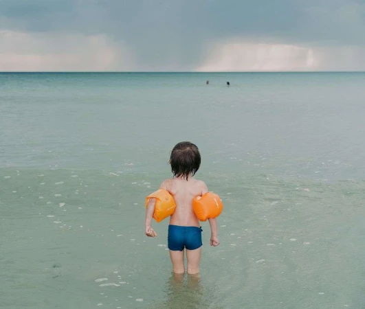 Young child wearing bright orange water wings and blue swim trunks standing in shallow ocean water
