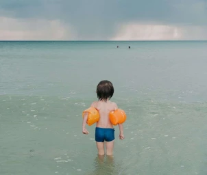 Young child wearing bright orange water wings and blue swim trunks standing in shallow ocean water