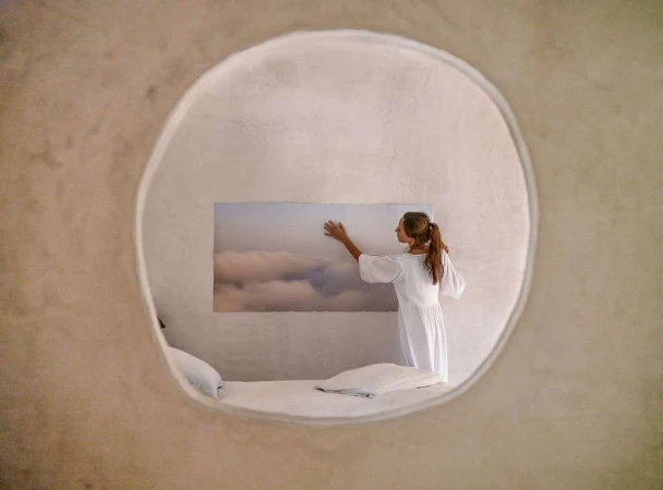 Woman in white dress touching a cloud artwork on a textured wall viewed through a large circular window opening
