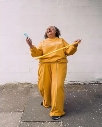 An elderly woman wearing a yellow tracksuit laughs while holding a smartphone and spinning a hula hoop outdoors.