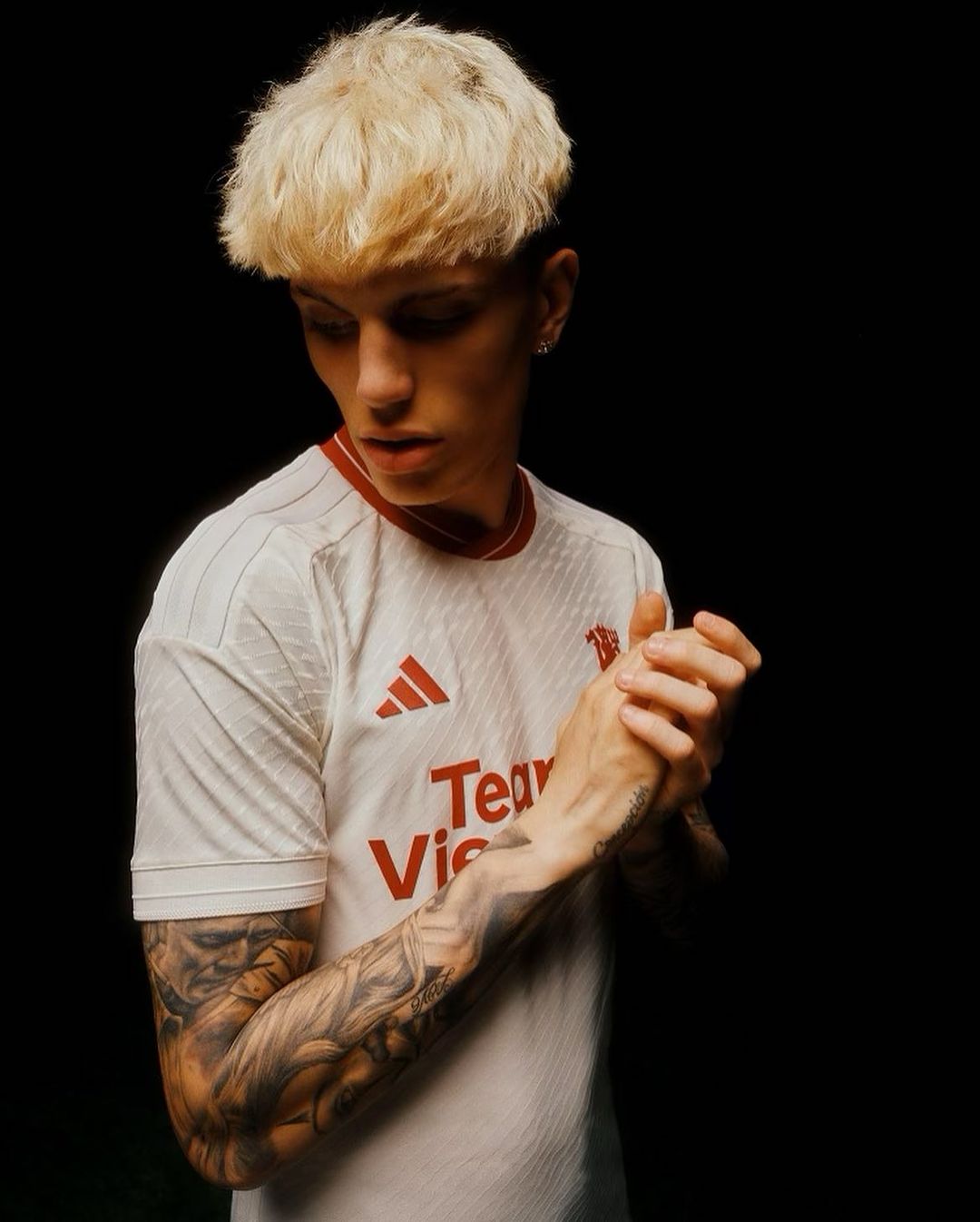 Portrait of a young adult male with platinum blond hair, fair skin, and tattoos wearing a white jersey against a dark backdrop.