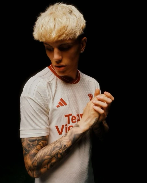 Portrait of a young adult male with platinum blond hair, fair skin, and tattoos wearing a white jersey against a dark backdrop.