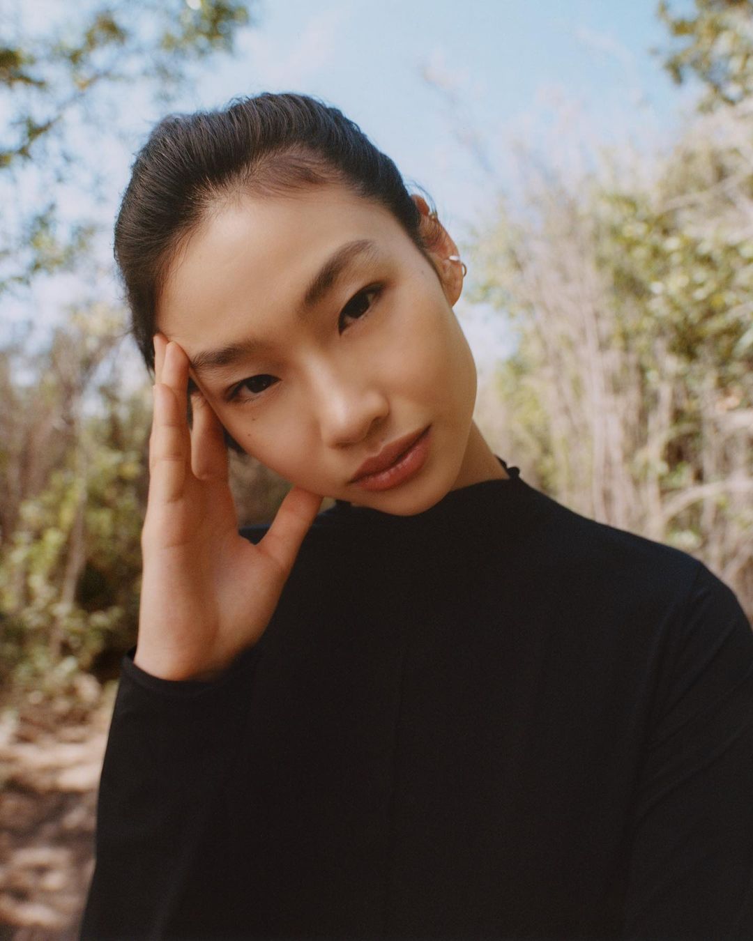 Close-up portrait of an Asian woman with her hand near her forehead, wearing a black top, outdoors in a sunny natural setting.