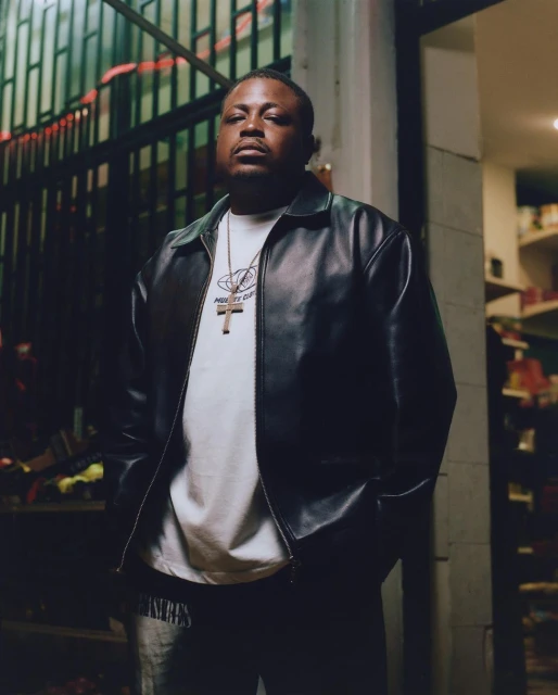 Studio shot of an adult male in a black leather jacket and white t-shirt, posing against a dark gated doorway. 