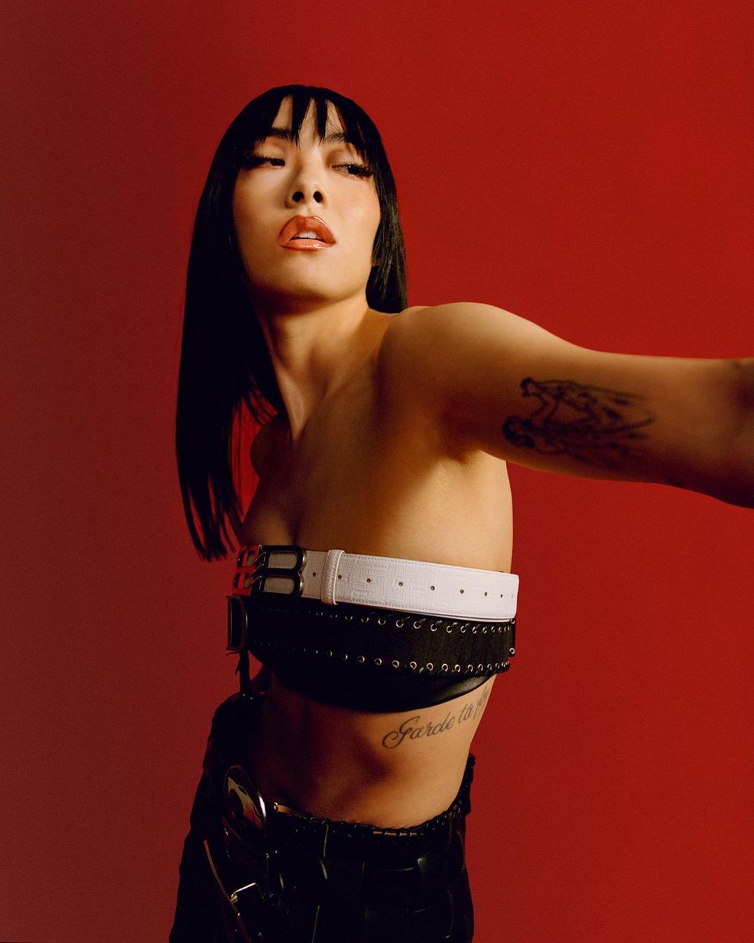 A stylish Asian woman with black hair in an unconventional belt top poses against a bold red backdrop, showcasing fashion-forward aesthetic.