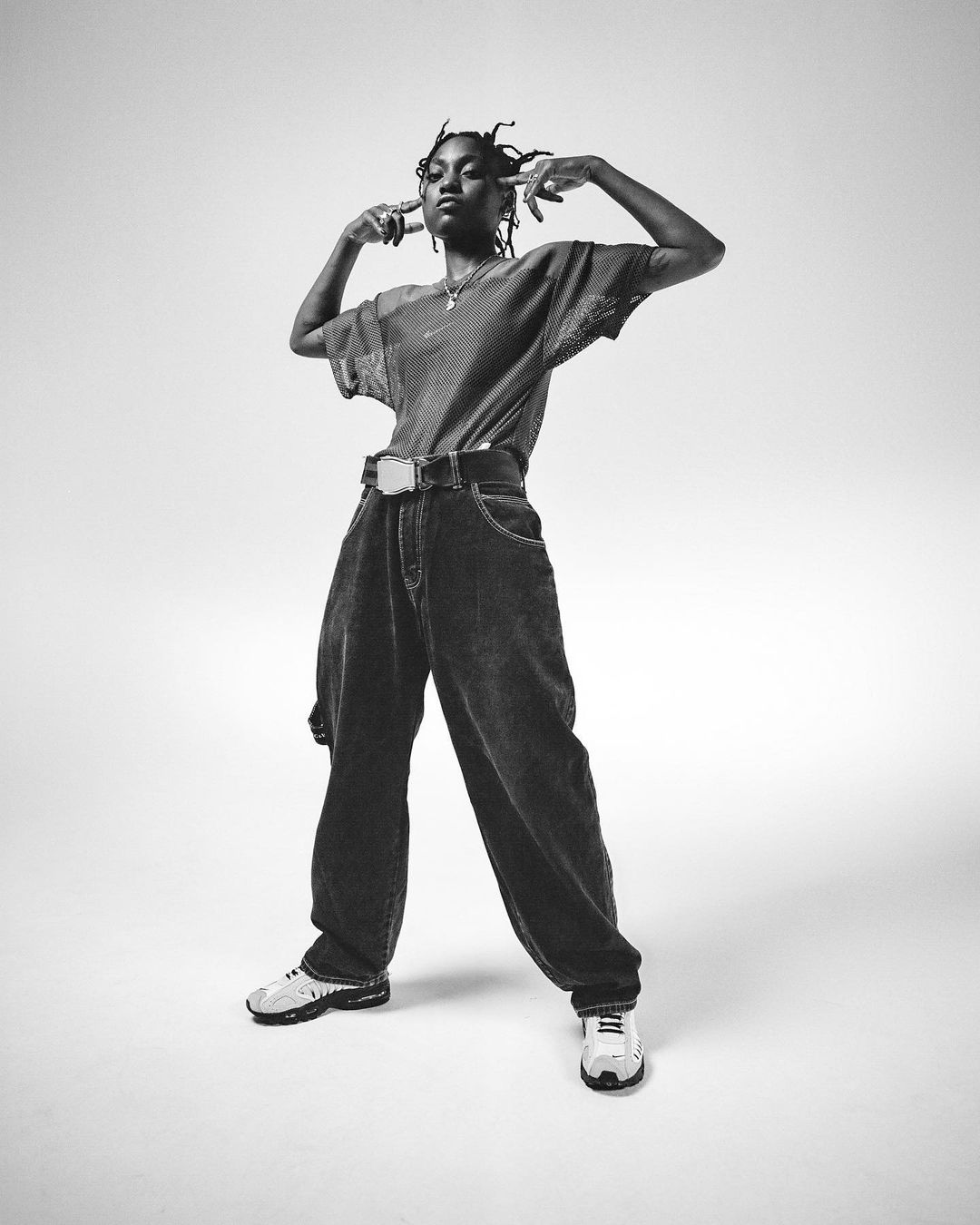 Black and white portrait of a slender young adult in loose denim, standing in a photo studio with a light neutral backdrop.