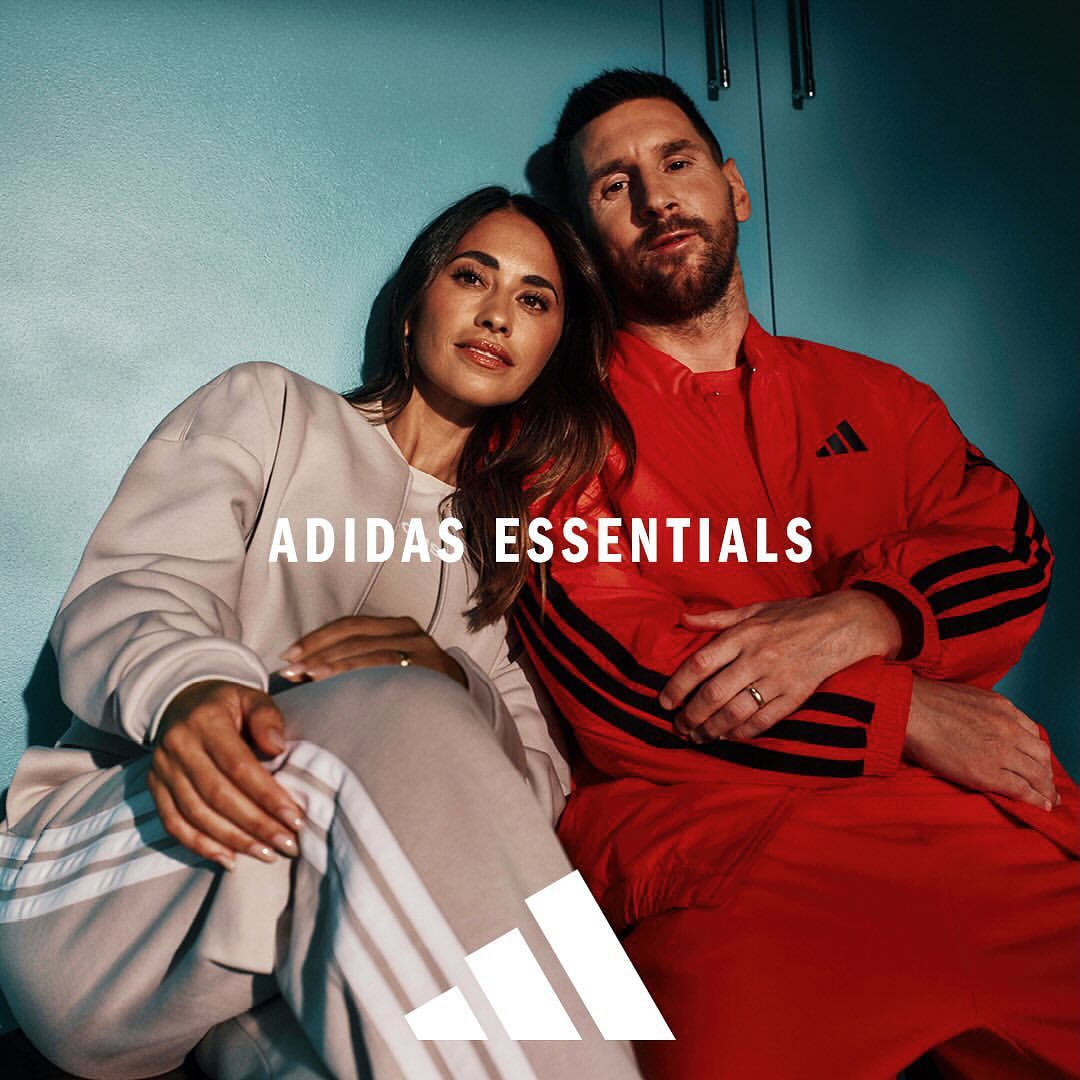 A smiling couple models Adidas Essentials loungewear in front of a blue-grey wall, showcasing coordinated comfort and style.