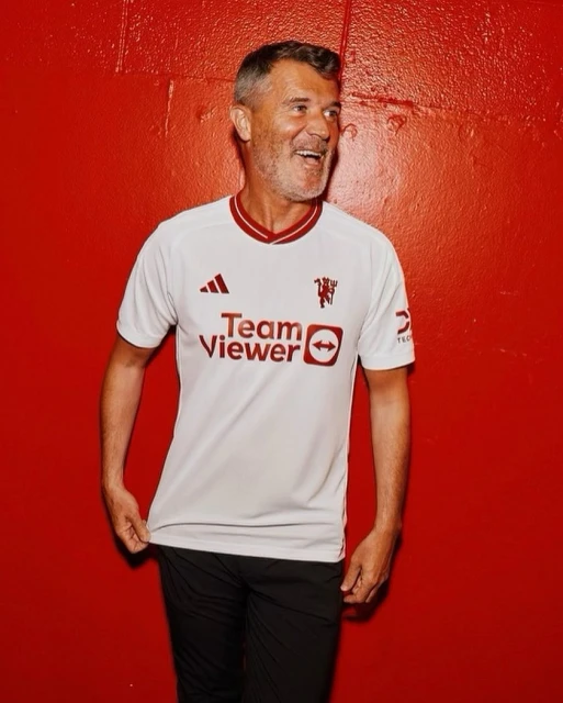 Adult man in a white and maroon Manchester United soccer jersey smiles in front of a red background.
