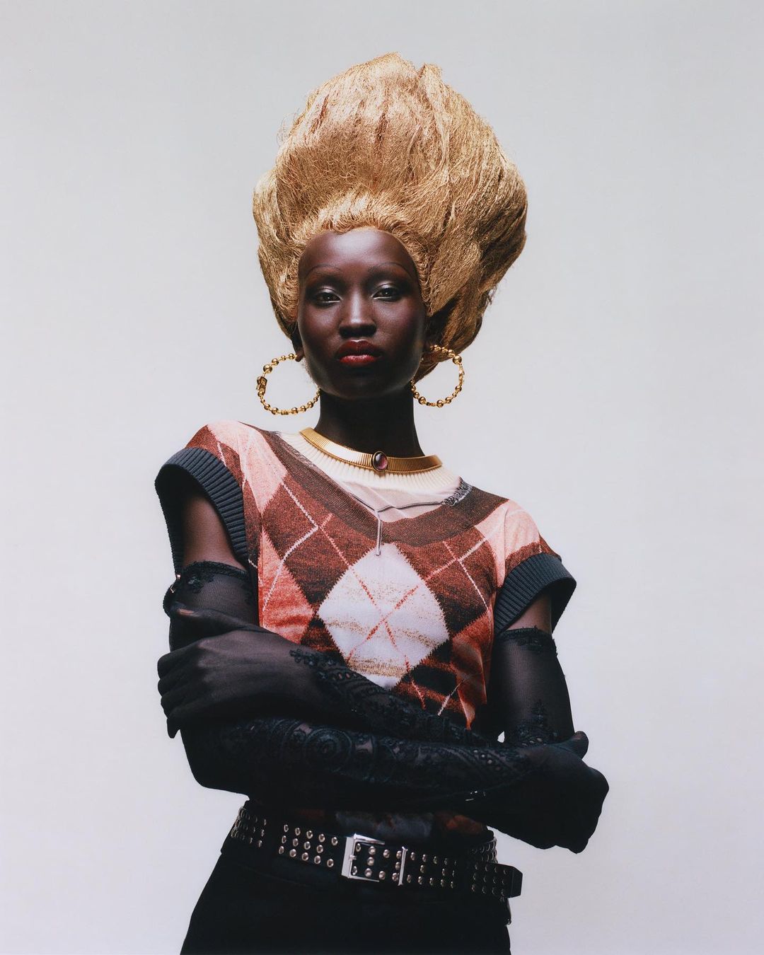 Fashion portrait of a woman with dark skin wearing gold earrings, a patterned sweater, and a large, elaborate blond hairstyle.