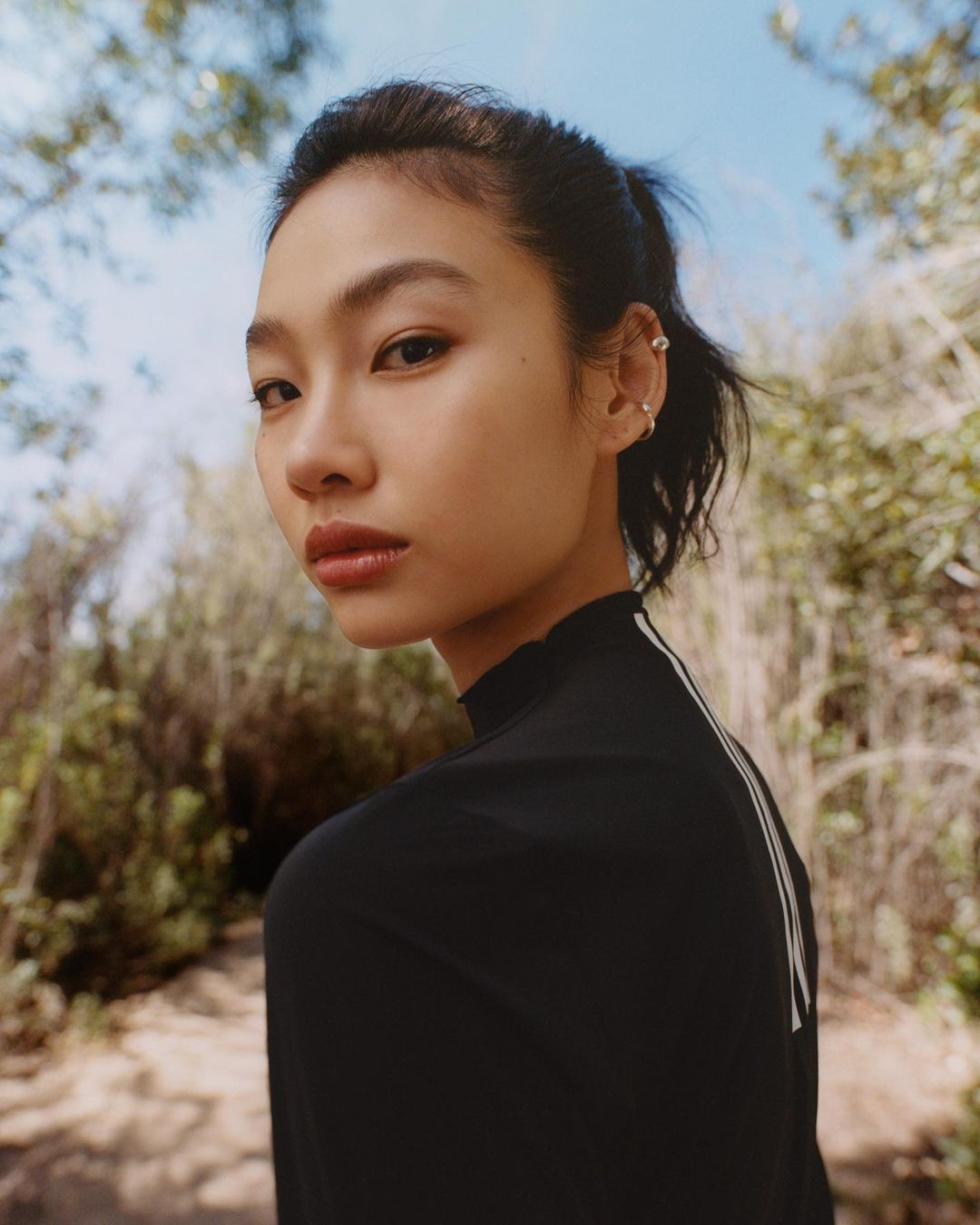 Portrait of a young Asian woman with dark hair pulled back, wearing a black top, posing outdoors with trees and sky in the background.