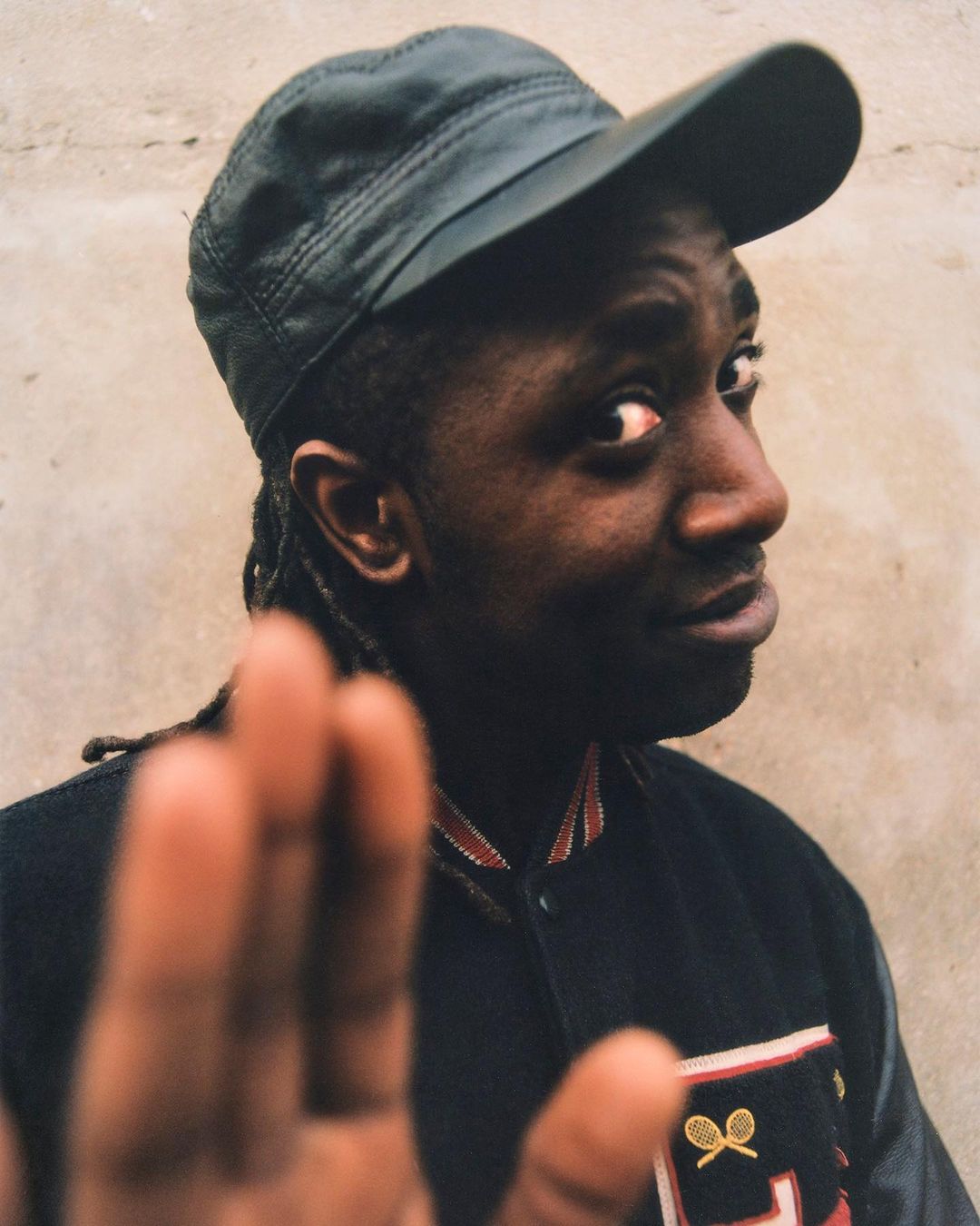 An adult male with an olive-green hat poses in an eye-level medium shot wearing a black jacket in front of a textured wall backdrop.