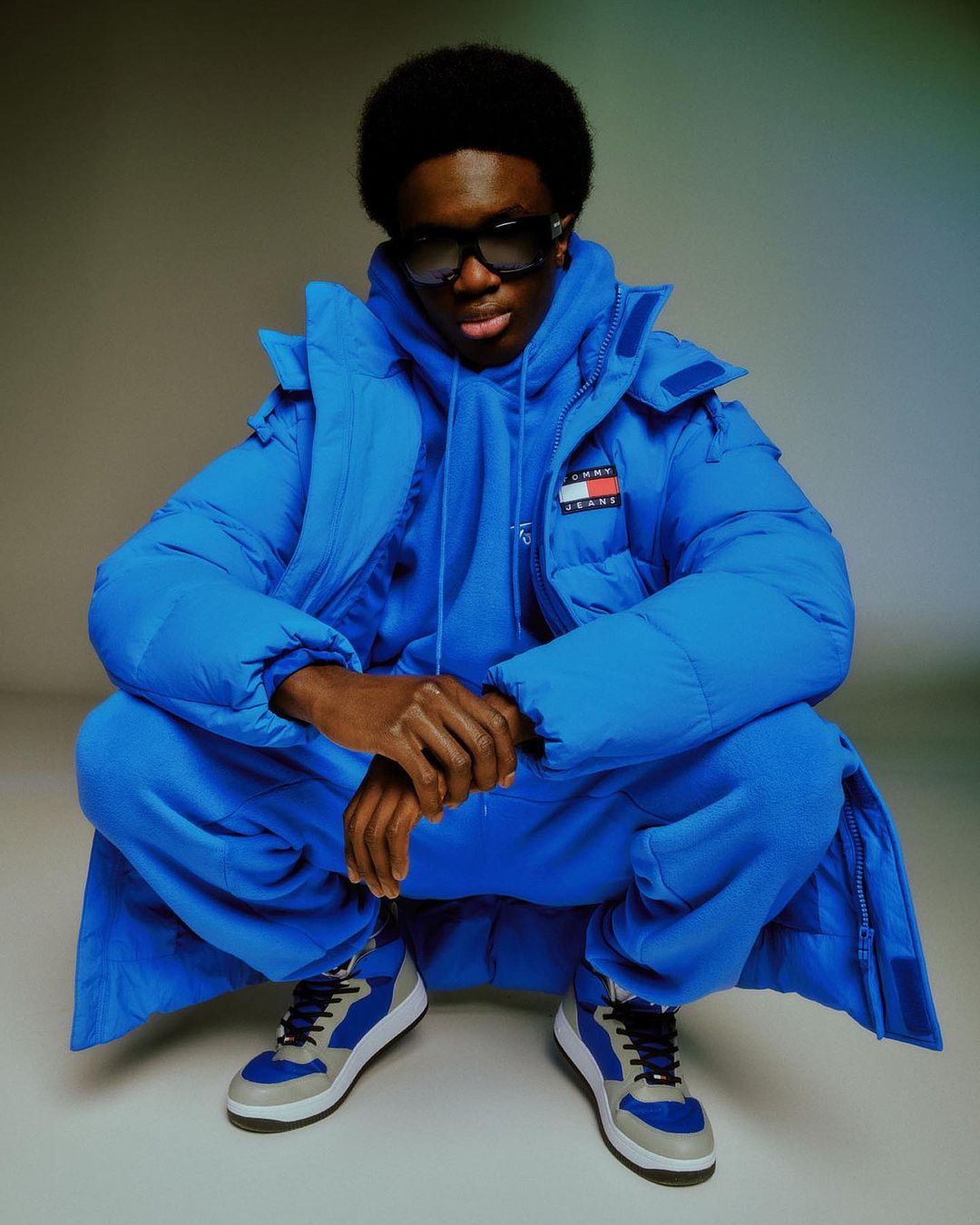 A young adult male with sunglasses in a blue down jacket and sweat suit crouches indoors in a fashion shot, highlighting cool tones.