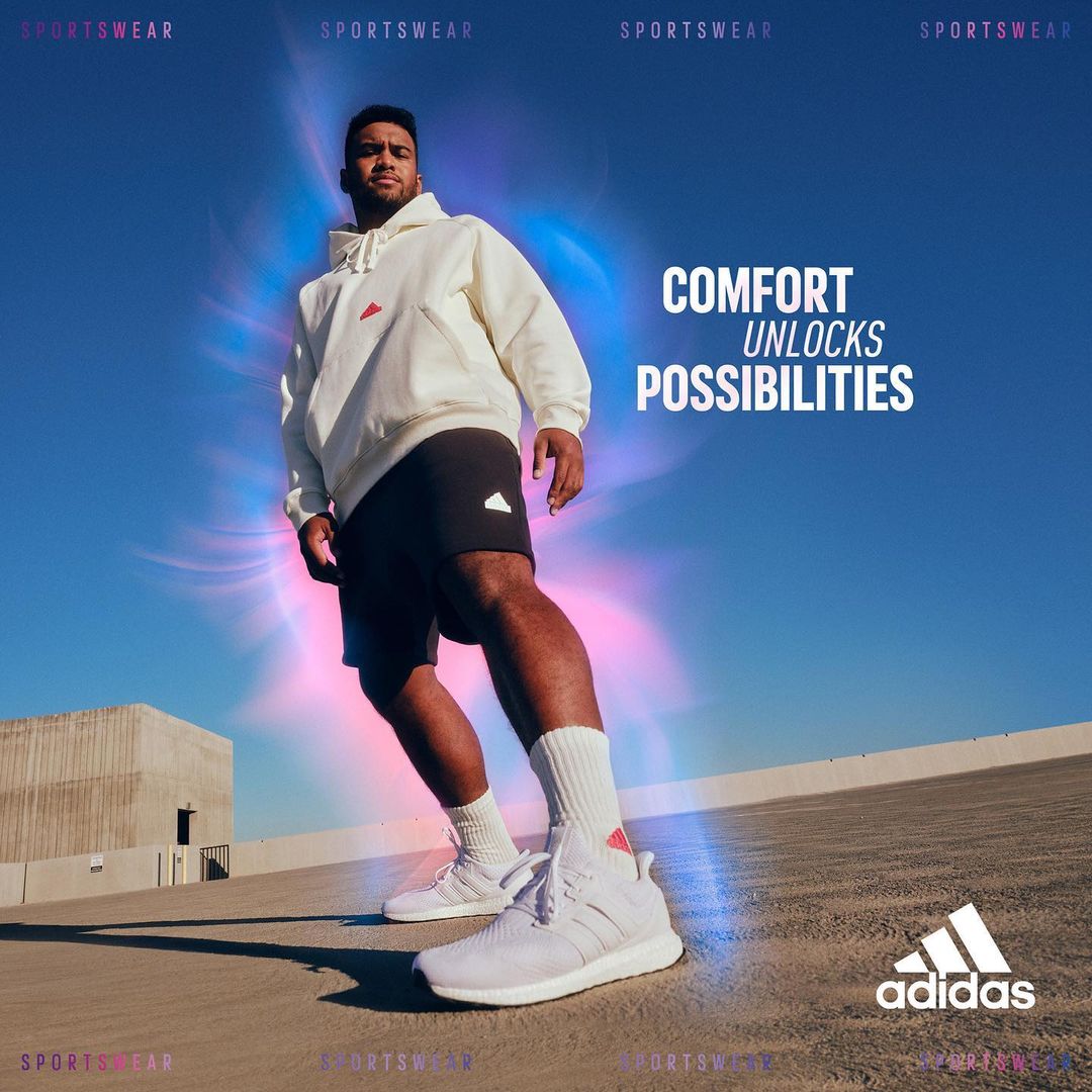 Male model in sportswear poses against a bright blue sky. He is wearing an off-white hoodie, black shorts, socks, and white athletic shoes.