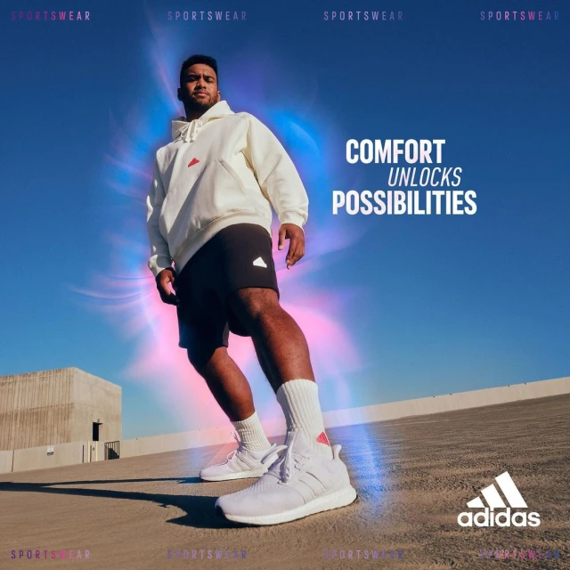 Male model in sportswear poses against a bright blue sky. He is wearing an off-white hoodie, black shorts, socks, and white athletic shoes.
