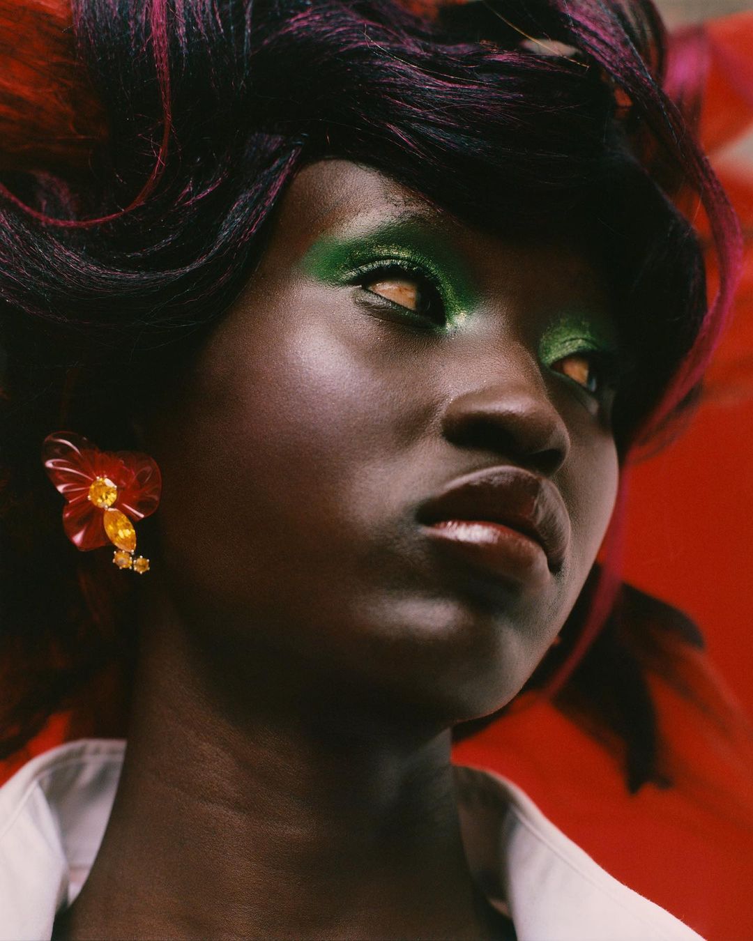 Close-up of a dark-skinned woman with colorful hair, bright green eye shadow, and an elaborate jeweled earring against red backdrop.