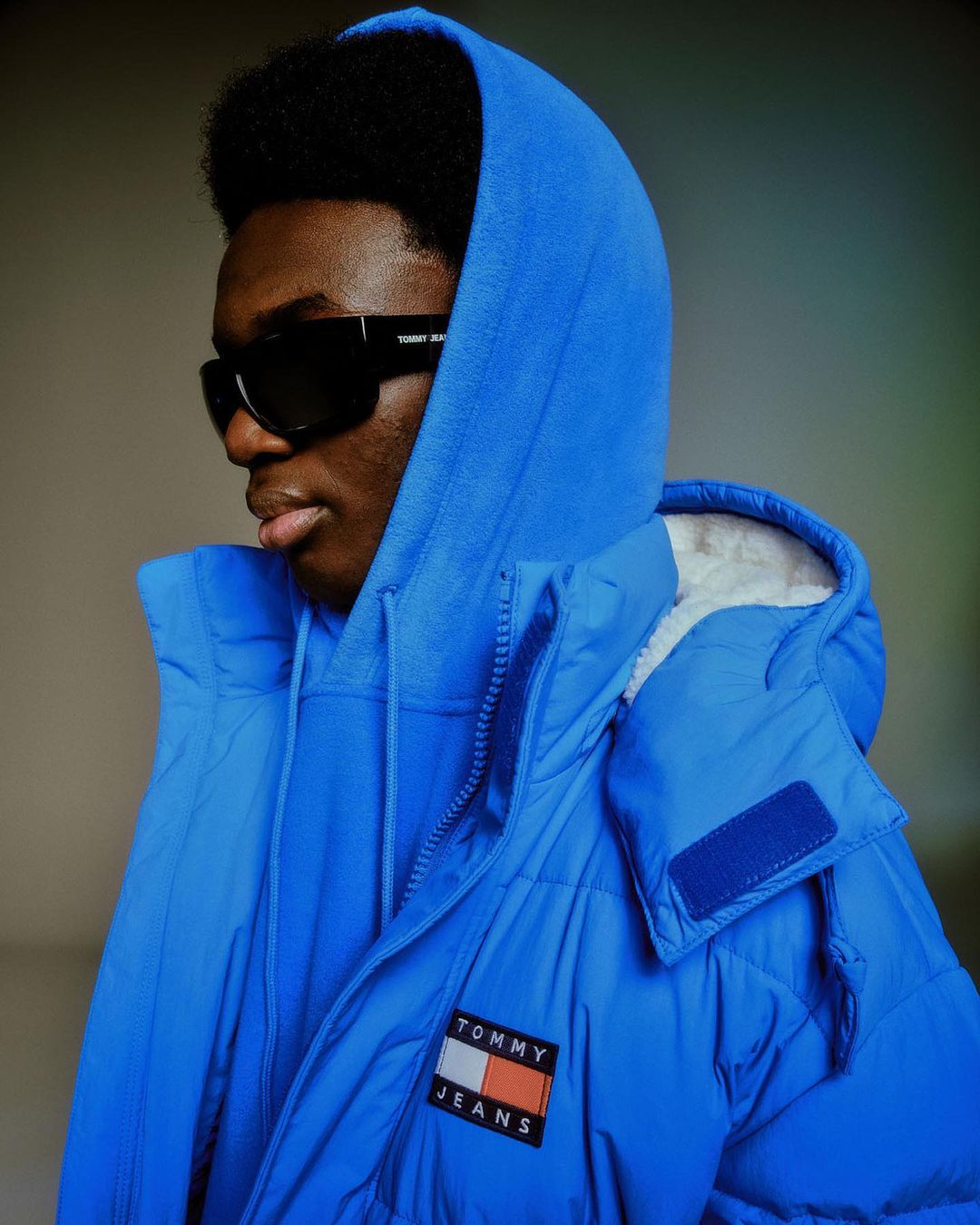 Headshot of a stylish Black male model wearing a blue puffer coat, matching hoodie, and black sunglasses posing indoors.