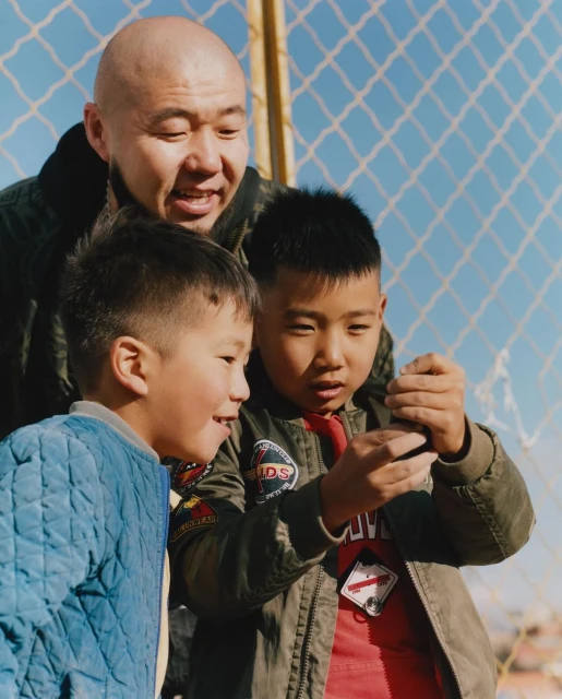 An adult Asian male and two Asian children standing outdoors in front of a chain-link fence, looking at a small object held by one child.