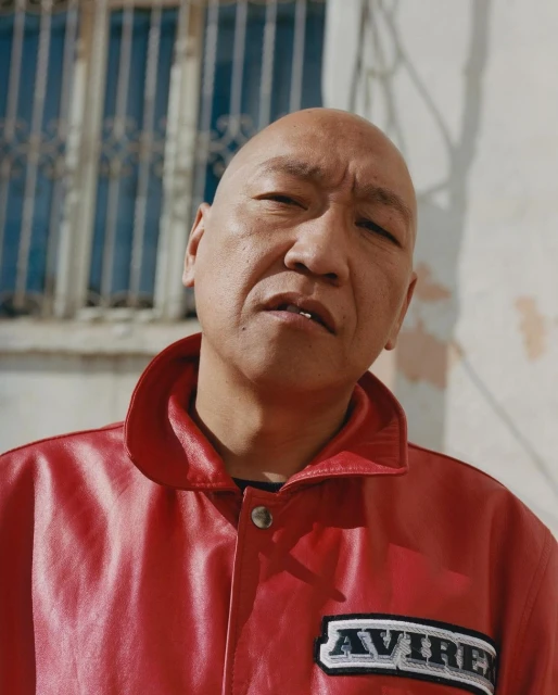 Portrait of a bald Asian adult male wearing a red leather jacket with an Avirex patch, posing outdoors near a window with metal bars.