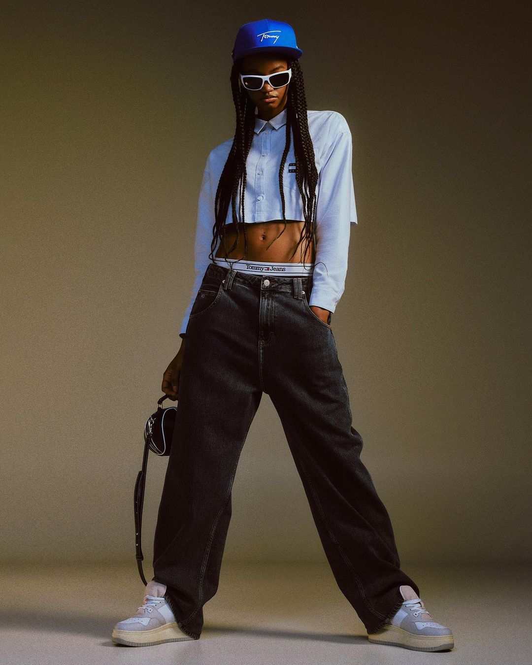 Full-length studio shot of a stylish adult with long braids wearing a blue baseball cap, sunglasses, crop top, and baggy jeans.