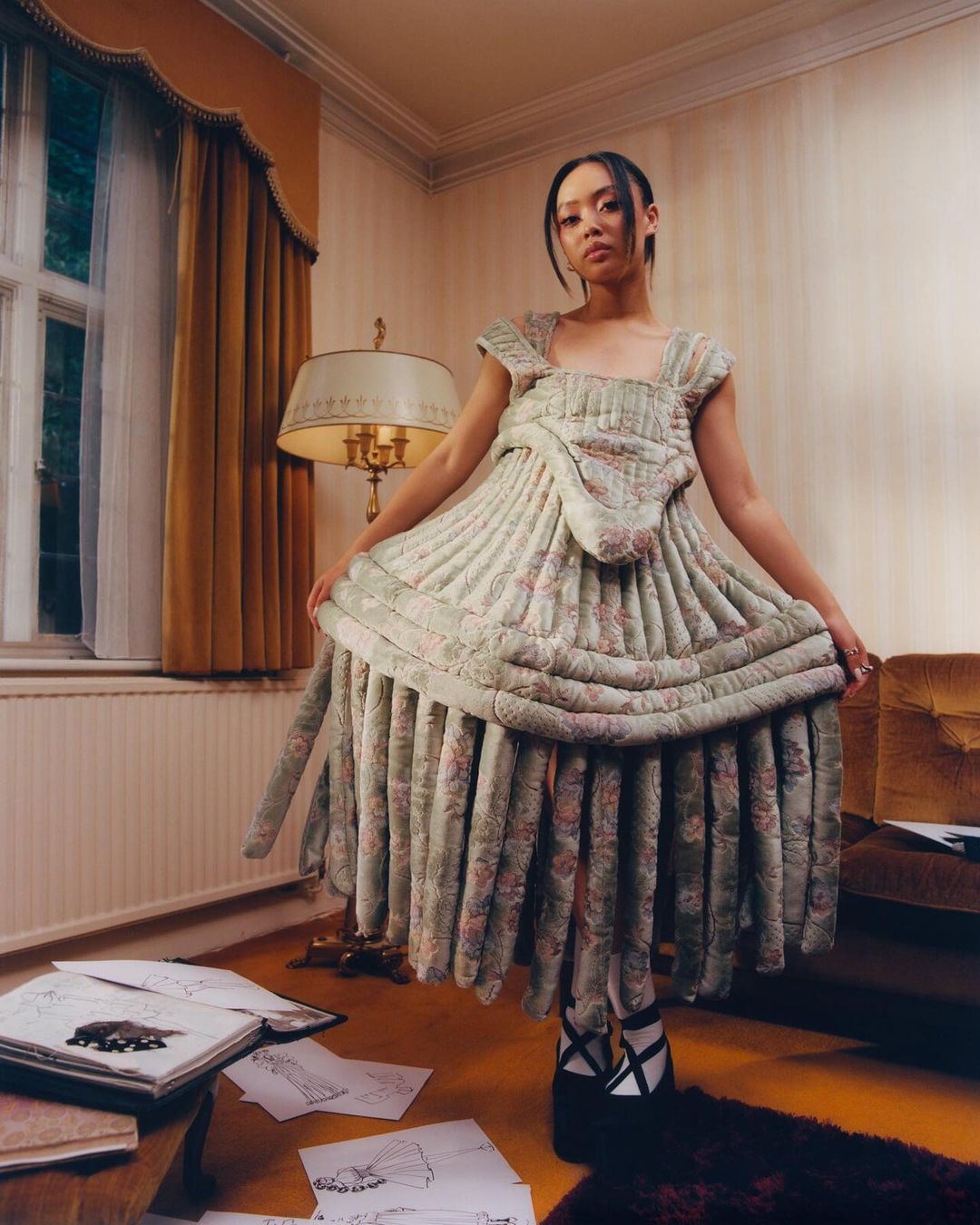 A woman poses indoors wearing a floral dress with drawings on the floor, creating a fashion editorial look in a vintage-styled interior.