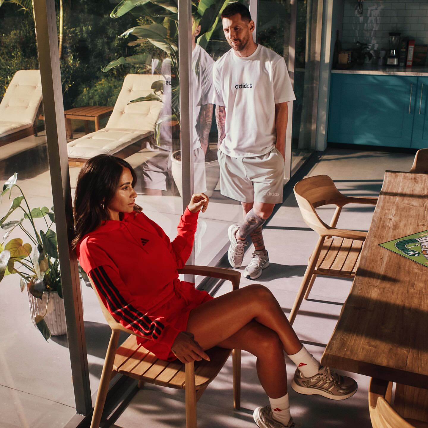 Two adults in athletic apparel pose indoors with natural light and modern decor in a fashion shot. A woman sits in a chair in red.