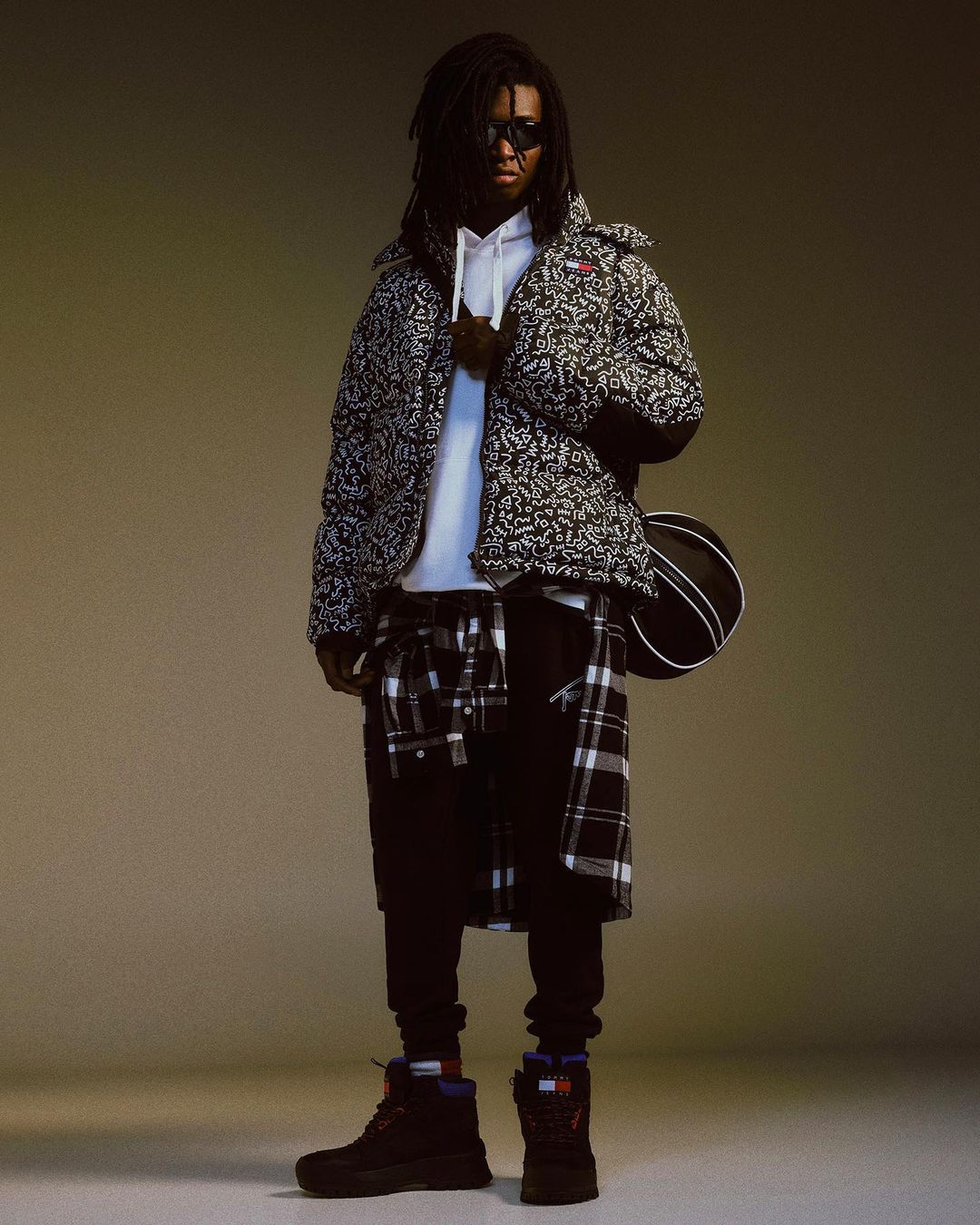 An adult male with dreadlocks wears a patterned puffer jacket, hoodie, and sunglasses, standing in front of a neutral studio backdrop.