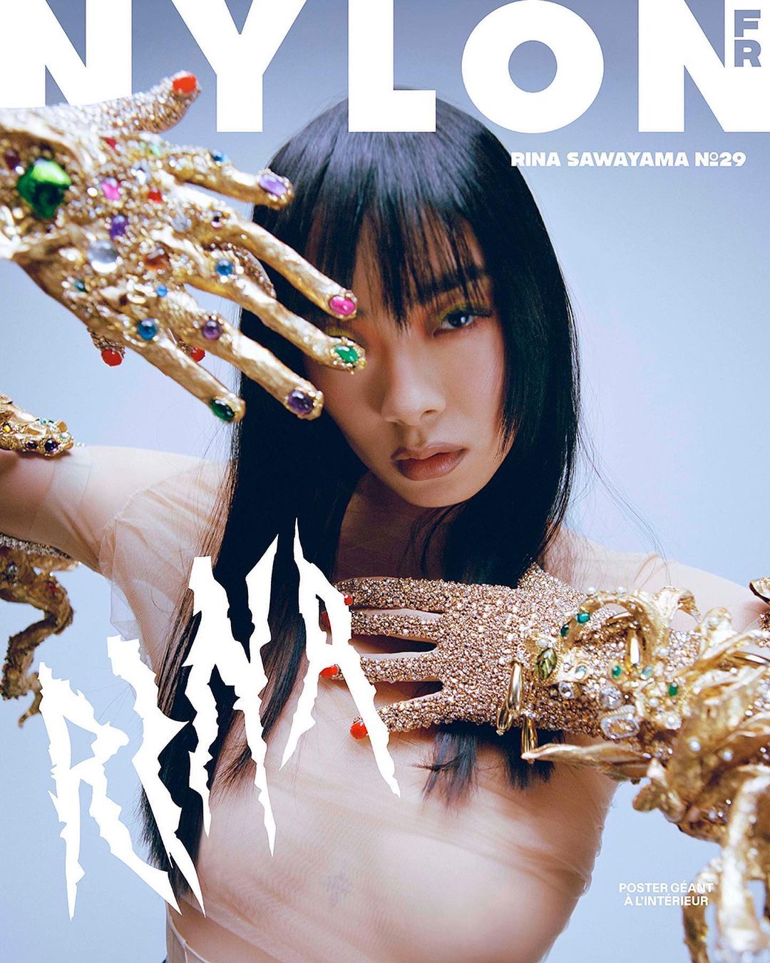 Magazine cover featuring Rina Sawayama with gold jeweled hands framing her face, black hair, and a pale blue background.