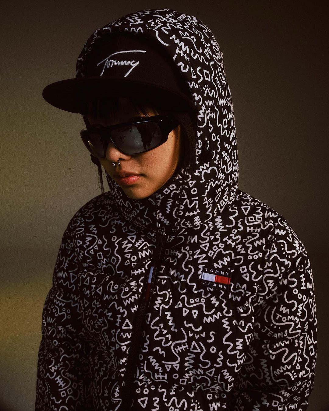 A young adult wearing a black patterned hooded jacket, sunglasses, and a baseball cap poses indoors in a close-up shot.