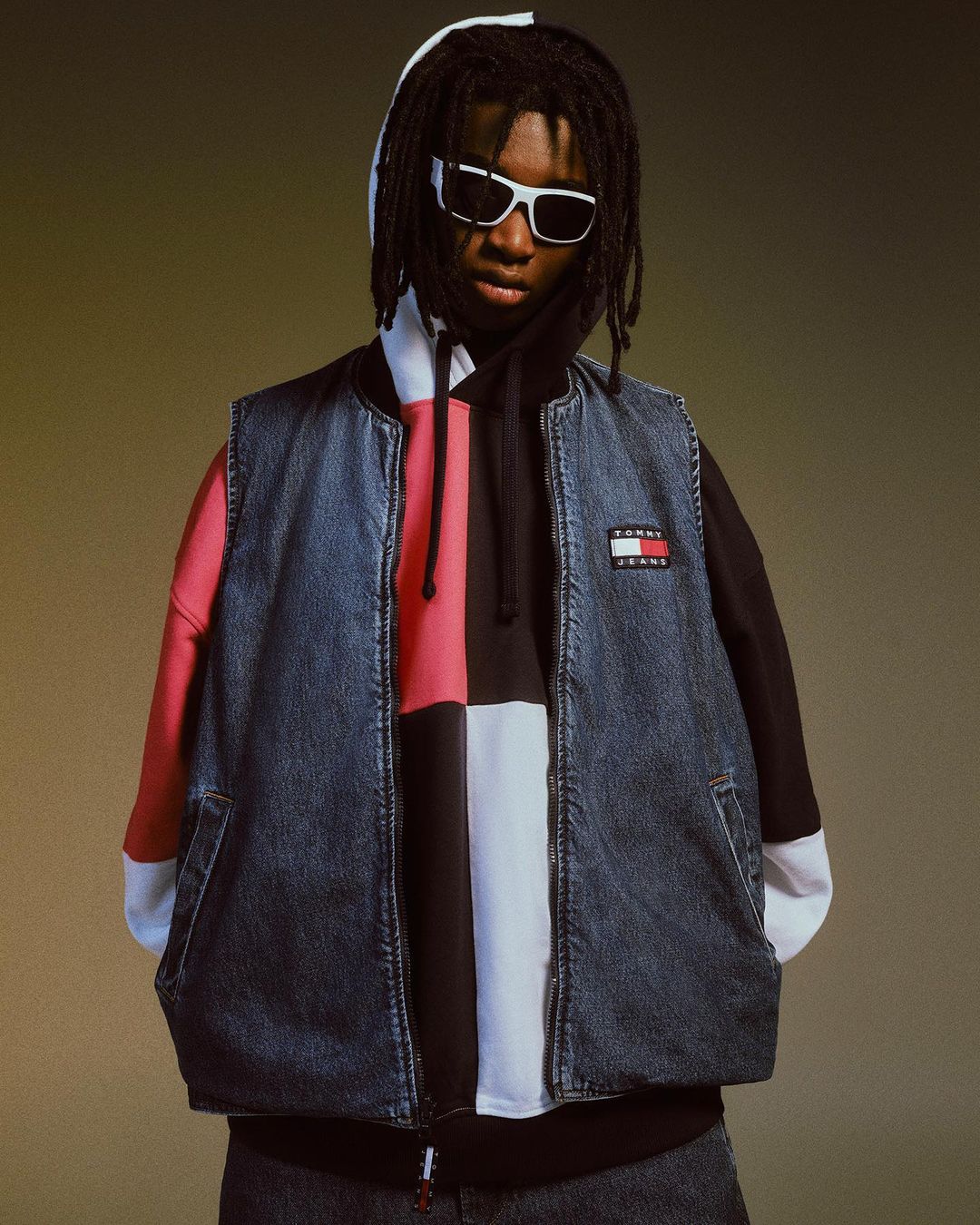 Young adult in denim vest over hoodie, wearing white-framed sunglasses, poses in a studio, showcasing urban fashion.