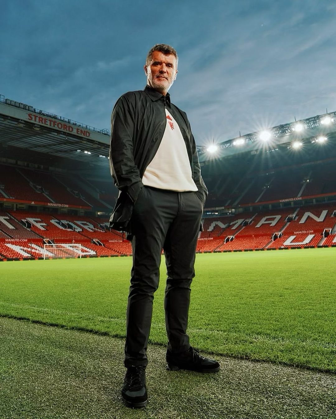 Full body shot of a male adult in black casual jacket and pants standing on a football field, stadium seating in the background.