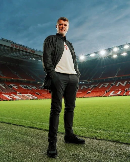 Full body shot of a male adult in black casual jacket and pants standing on a football field, stadium seating in the background.