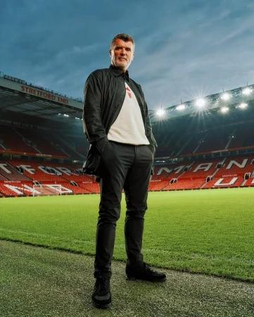 Full body shot of a male adult in black casual jacket and pants standing on a football field, stadium seating in the background.