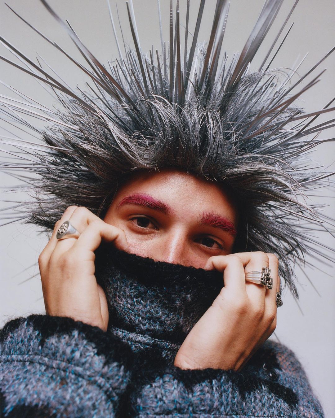 Close-up portrait of a young adult with spiky silver hair and eyebrows dyed bright pink, wearing a grey knit turtleneck sweater.