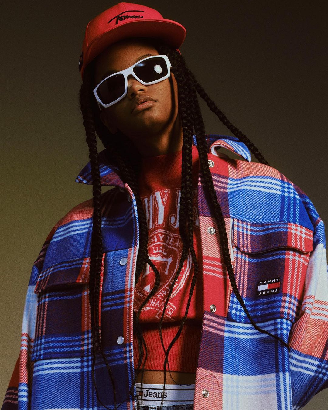 Woman wearing a Tommy Jeans red cap and plaid jacket poses. She wears white sunglasses and stands in front of a solid neutral backdrop.
