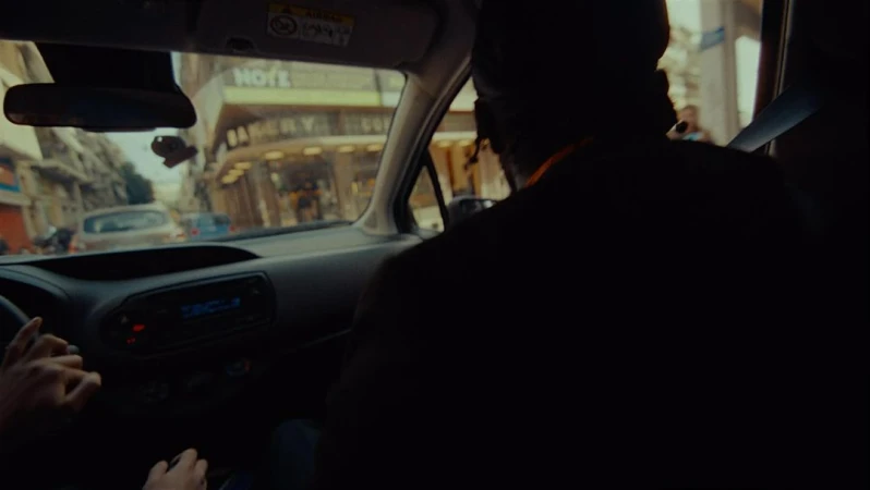 Interior view from a car with two people in the front seats and a person in the back with buildings visible through the windshield.