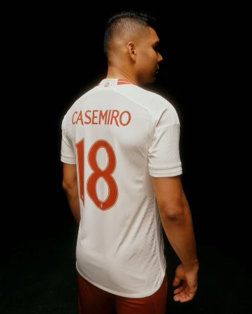 Soccer player Casemiro in a white jersey with red number 18, set against a dark background. Profile view focusing on uniform details.