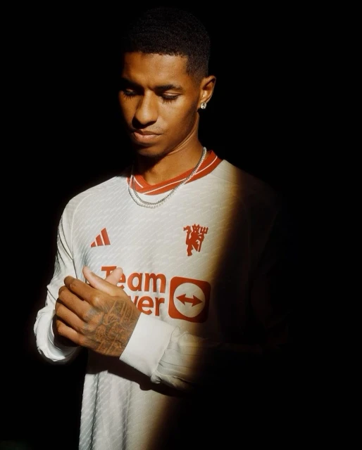 Adult male with short dark hair, wearing a white and red long-sleeved Manchester United soccer jersey, stands in a dark indoor setting.
